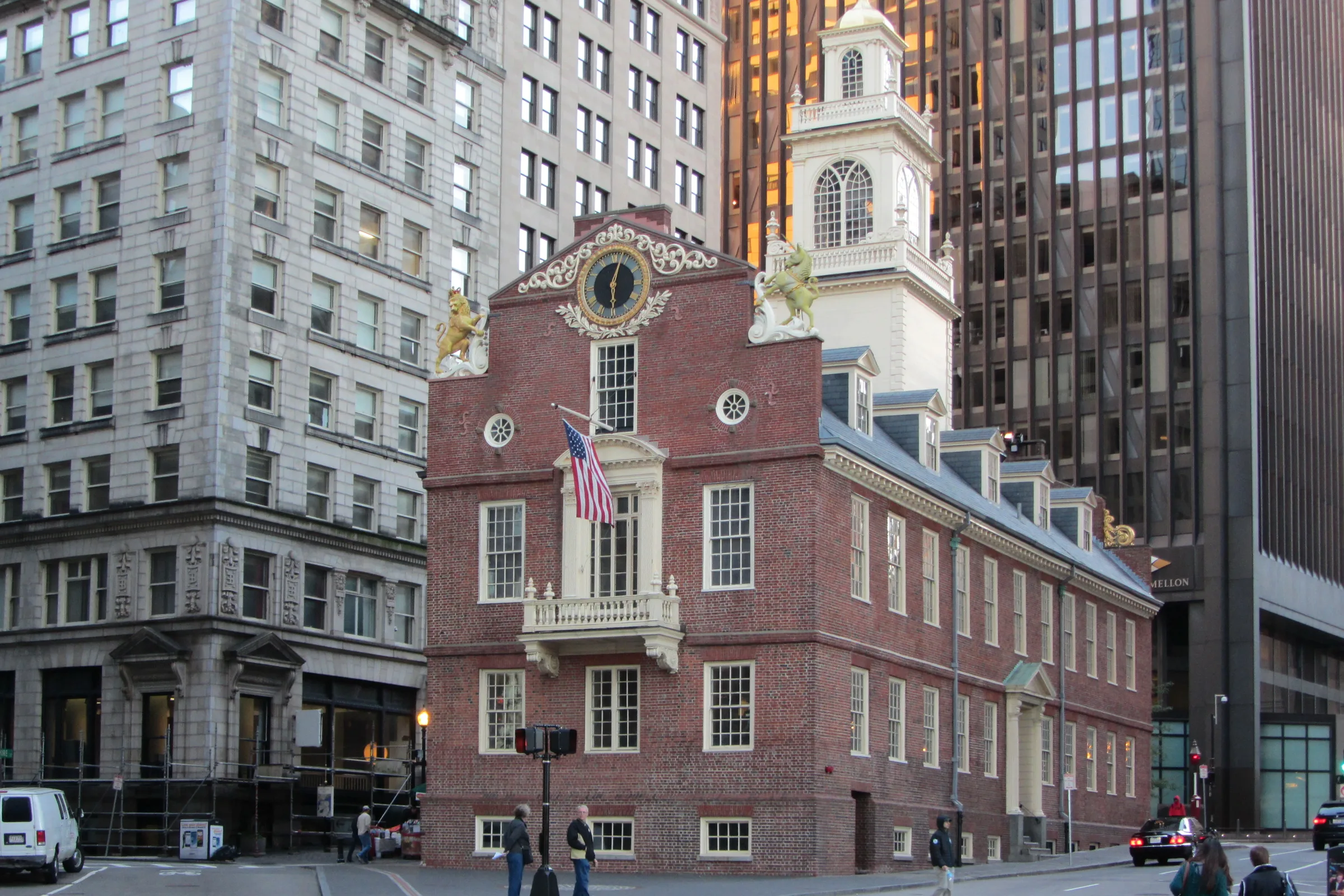 The Old State House in present-day Boston, its colonial brick façade and clock tower hemmed in by surrounding glass and stone office towers at the corner of Washington and State Streets.